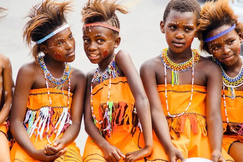 Eine Gruppe kleiner Kinder aus Südafrika sitzt zusammen und trägt leuchtend orangefarbene, mit Perlen besetzte traditionelle Kleidung und weiße Gesichtsbemalung.