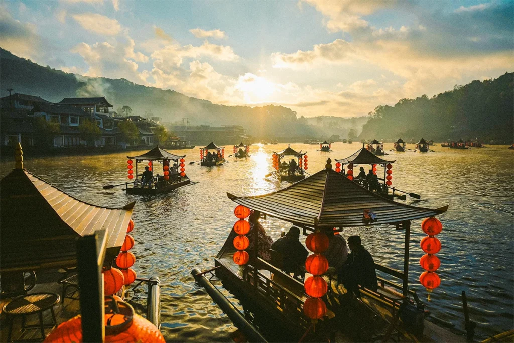 Mehrere traditionelle Boote mit roten Laternen und Menschen darauf, die bei Sonnenuntergang auf einem See vor einer Bergkulisse in Thailand fahren.