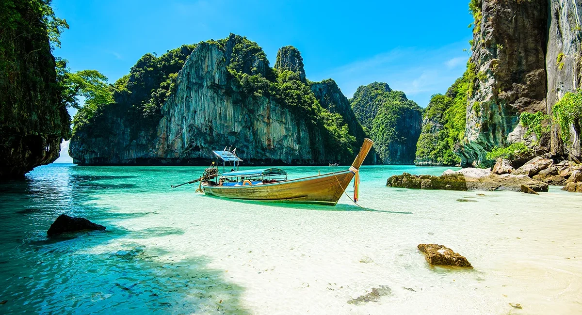 Eine tropische Bucht in Thailand mit kristallklarem türkisfarbenem Wasser, weißem Sand, dramatischen Kalksteinfelsen und einem traditionellen Langheckboot.