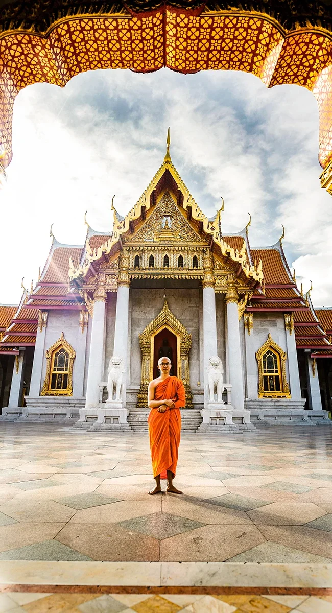 Ein Mönch in orangefarbenem Gewand steht vor dem weißen thailändischen Mamortempel mit Goldelementen.