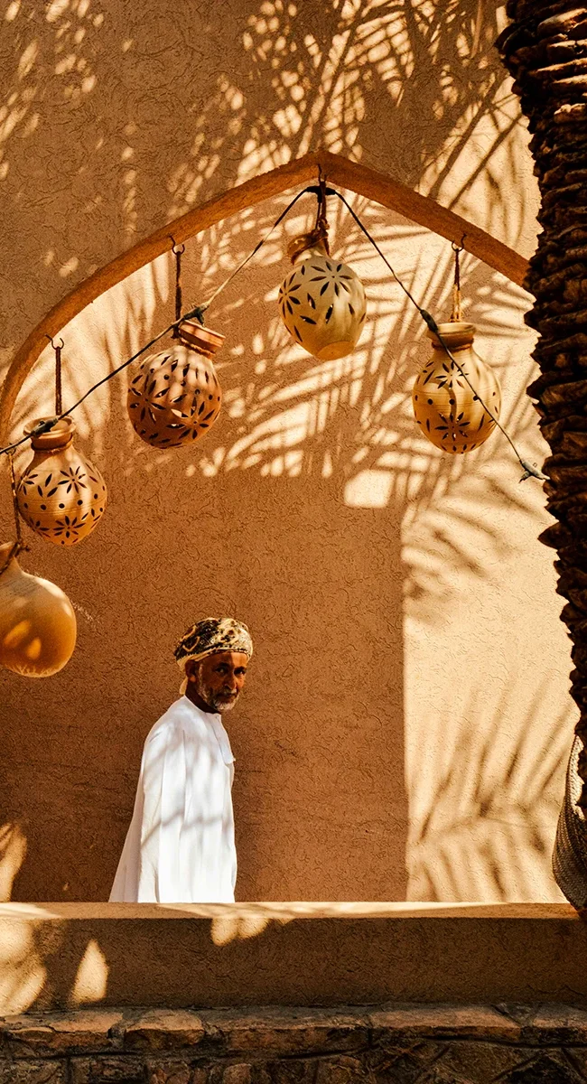 Ein arabischer Mann in traditionellem weißen Gewand geht hinter einem Bogen entlang, über dem Tonkannen hängen, während Palmenschatten die Szene überlagert.