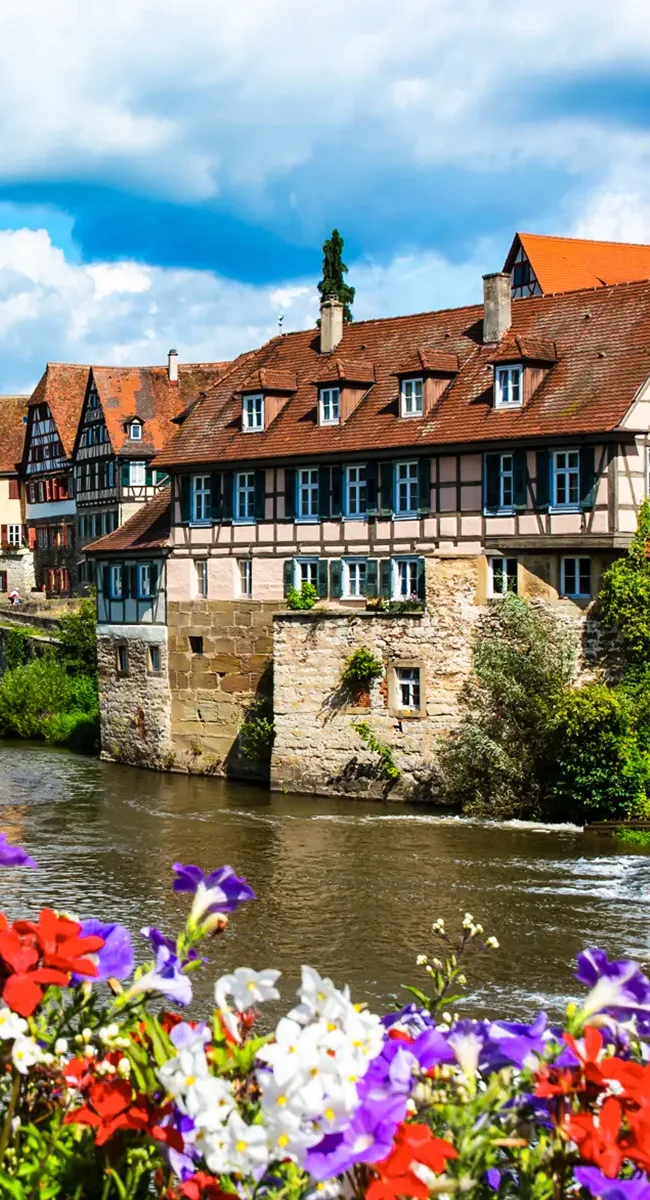 Der historische Keckenhof mit seinen Fachwerkhäusern direkt am Fluss Kocher in Schwäbisch Hall, eingerahmt von bunten Blumen im Vordergrund.