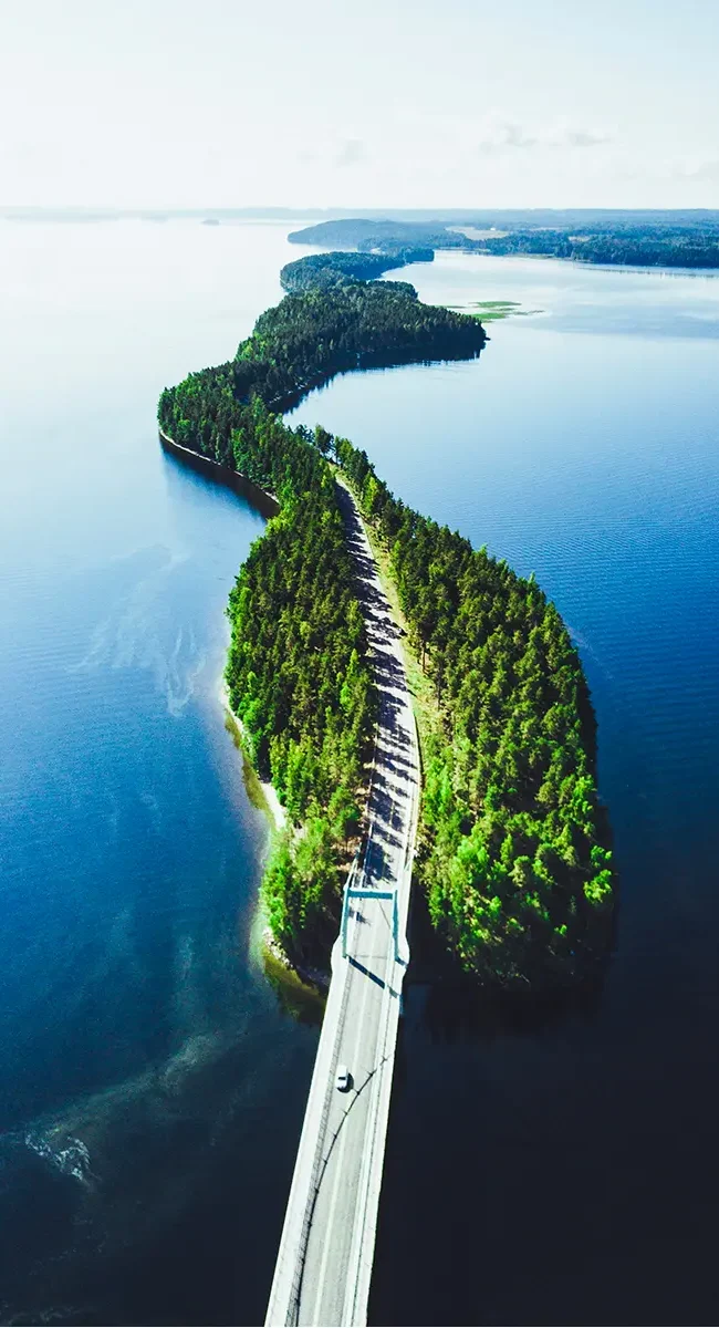 Luftaufnahme einer Straße zwischen grünem Sommerwald und über einen See in Finnland