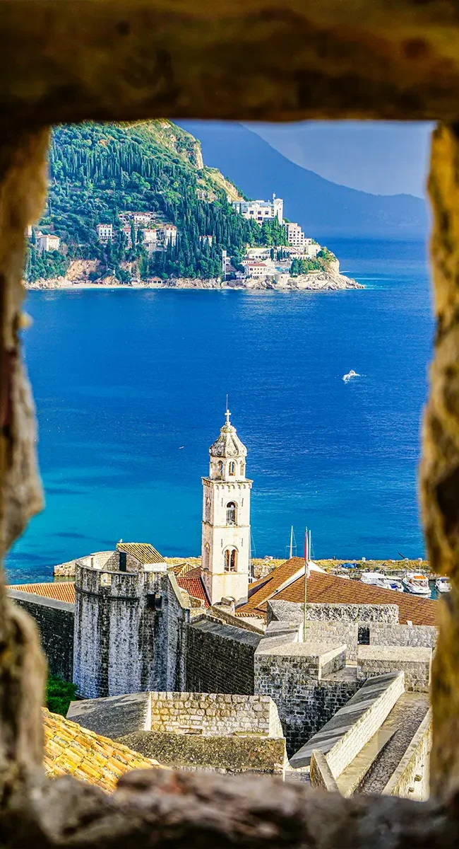 Blick durch eine steinerne Öffnung in der Altstadtmauer von Dubrovnik auf einen markanten Kirchturm und das dahinter liegende Meer.