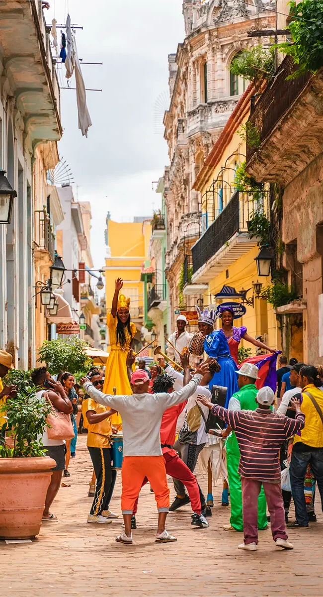 Eine belebte Straße in Havanna, Kuba, ist gefüllt mit Menschen, Musikern und farbenfrohen Stelzenläufern während einer lebhaften Parade oder eines Festivals.