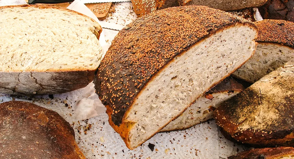 Mehrere lettische rustikale, dunkle Brote und angeschnittene Brotscheiben, die mit Samen wie Sesam bestreut sind und auf einer weißen Unterlage liegen.