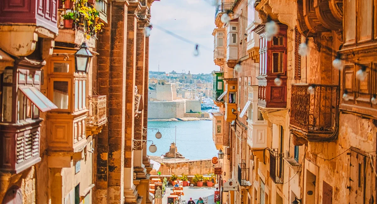 Eine steile, enge Straße in Valletta, Malta, gesäumt von Gebäuden mit bunten Holzbalkonen, die einen malerischen Blick auf den Großen Hafen im Hintergrund bietet.