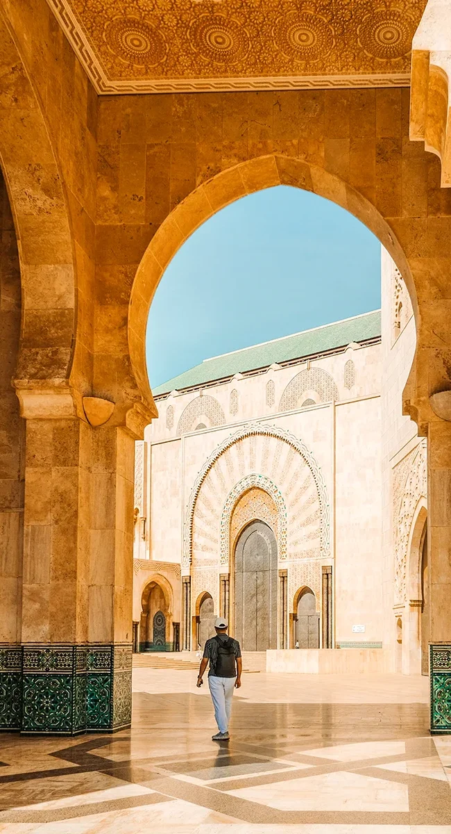 Eine Person spaziert unter großen, reich verzierten Steinbögen auf den Innenhof einer Moschee in Marokko zu, während die Sonne Muster auf den Marmorboden wirft.