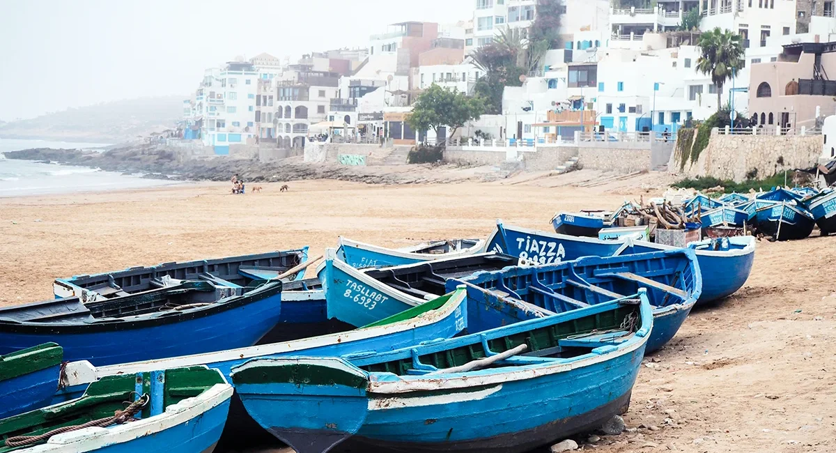 Am Strand eines Küstenortes in Marokko liegen mehrere traditionelle, blau bemalte Fischerboote im Sand vor einer Kulisse aus weißen Gebäuden am Hang.