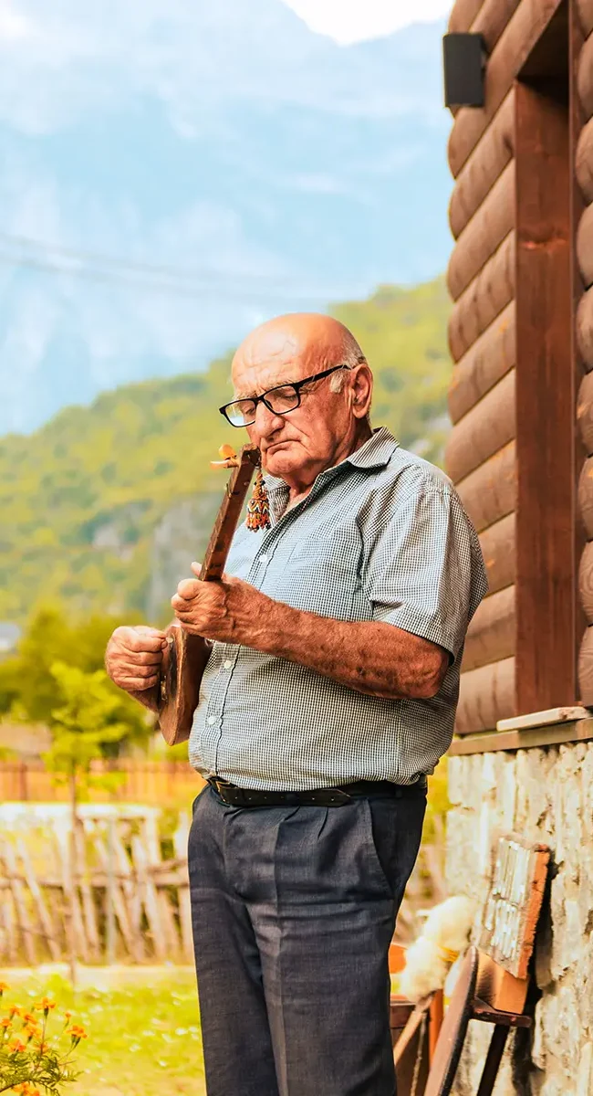 Ein älterer einheimischer Mann steht vor einer Holzhütte in bergiger Landschaft und spielt ein traditionelles Musikinstrument aus Montenegro.
