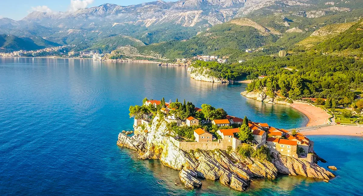 Die kleine Insel Sveti Stefan in Montenegro, die durch einen schmalen Damm mit dem Festland verbunden ist und von klarem, blauem Wasser sowie Bergen umgeben wird.