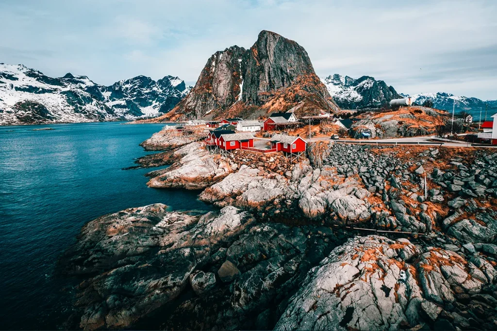 Ein Fischerdorf auf den norwegischen Lofoten-Inseln, mit traditionellen roten Fischerhütten an der felsigen Küste, umgeben von tiefblauem Wasser und steil aufragenden, schneebedeckten Bergen.