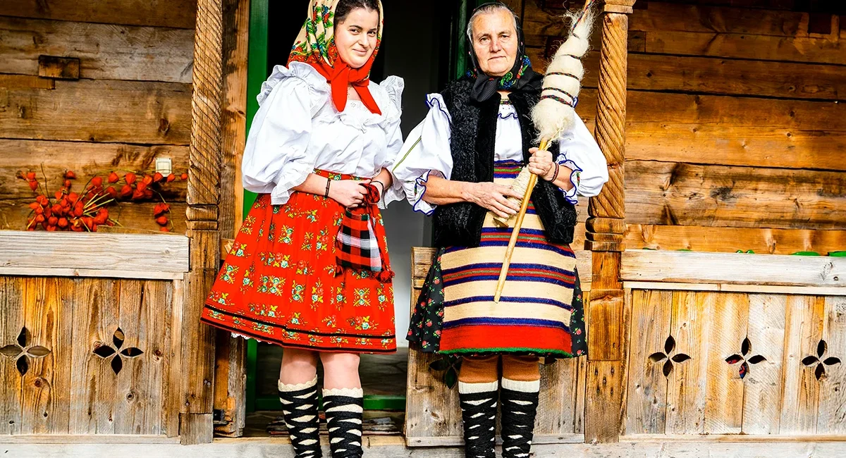 Zwei rumänische Bäuerinnen stehen vor einer Holzhütte und tragen traditionelle bunte Gewänder und Kopftücher.