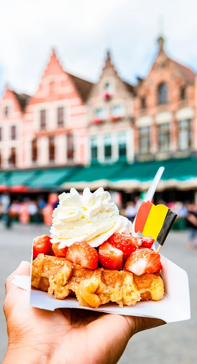 Auf einem Marktplatz mit bunten Häusern wird eine belgische Waffel mit Erdbeeren und Sahne gehalten.