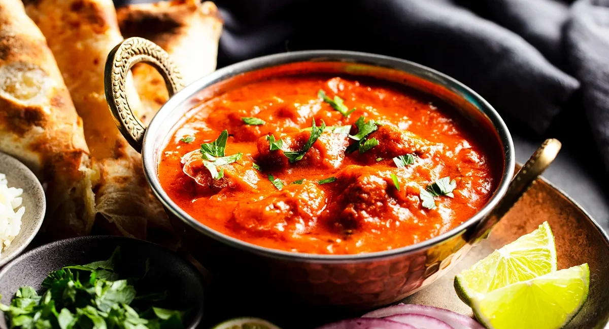 Ein indisches rotes Curry in einer Schüssel mit Limetten, Zwiebeln und Brot.