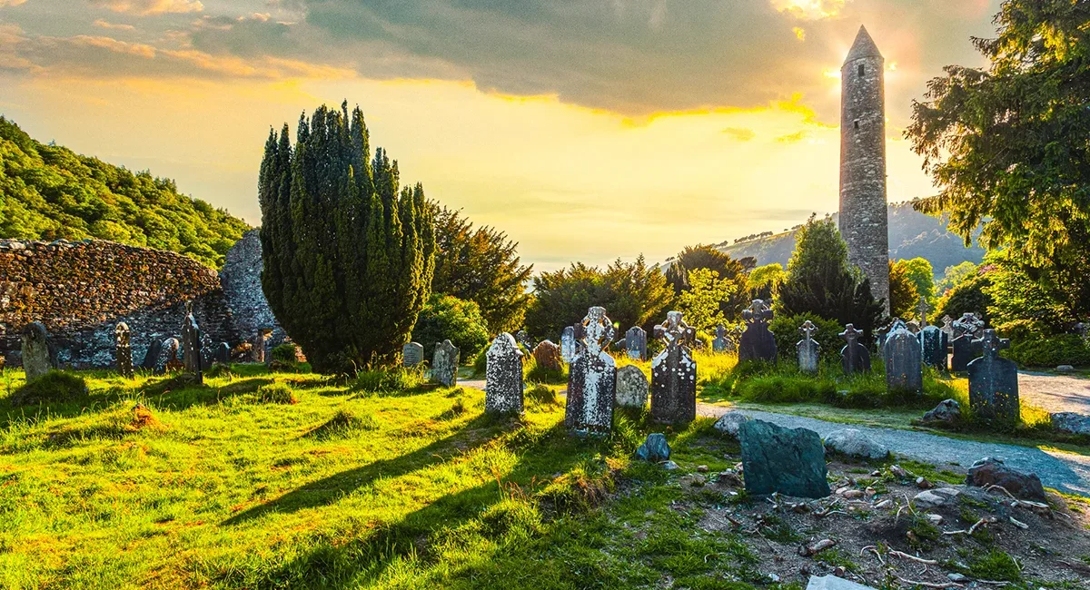 Der historische Rundturm und der alte Friedhof der mittelalterlichen Klostersiedlung Glendalough in Irland während eines stimmungsvollen Sonnenuntergangs.