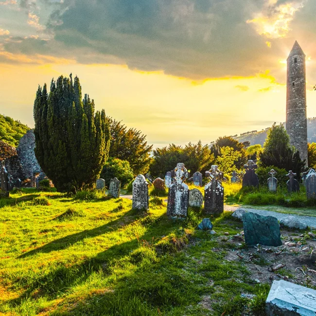 Der historische Rundturm und der alte Friedhof der mittelalterlichen Klostersiedlung Glendalough in Irland während eines stimmungsvollen Sonnenuntergangs.