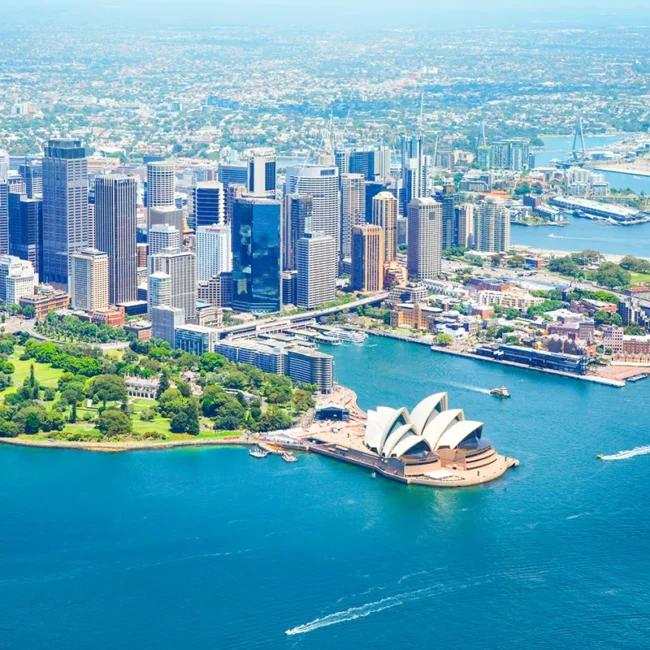 Luftaufnahme von Sydney bei Tageslicht: Das Opernhaus mit seinen weißen, segelförmigen Dächern liegt am Hafen neben der Harbour Bridge. Umgeben von blauem Wasser, Booten und einer dichten Skyline moderner Hochhäuser