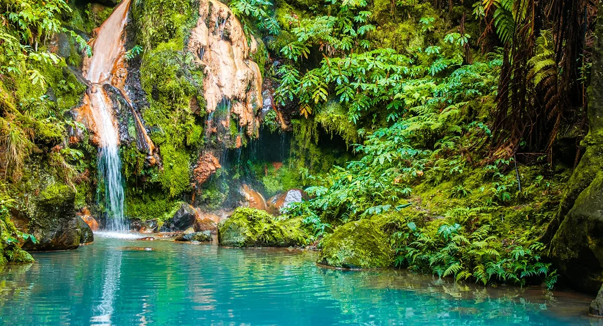 Ein Wasserfall im Naturpark Caldeira Velha auf der Azoreninsel São Miguel fließt zwischen dichter grüner Vegetation in ein Becken.