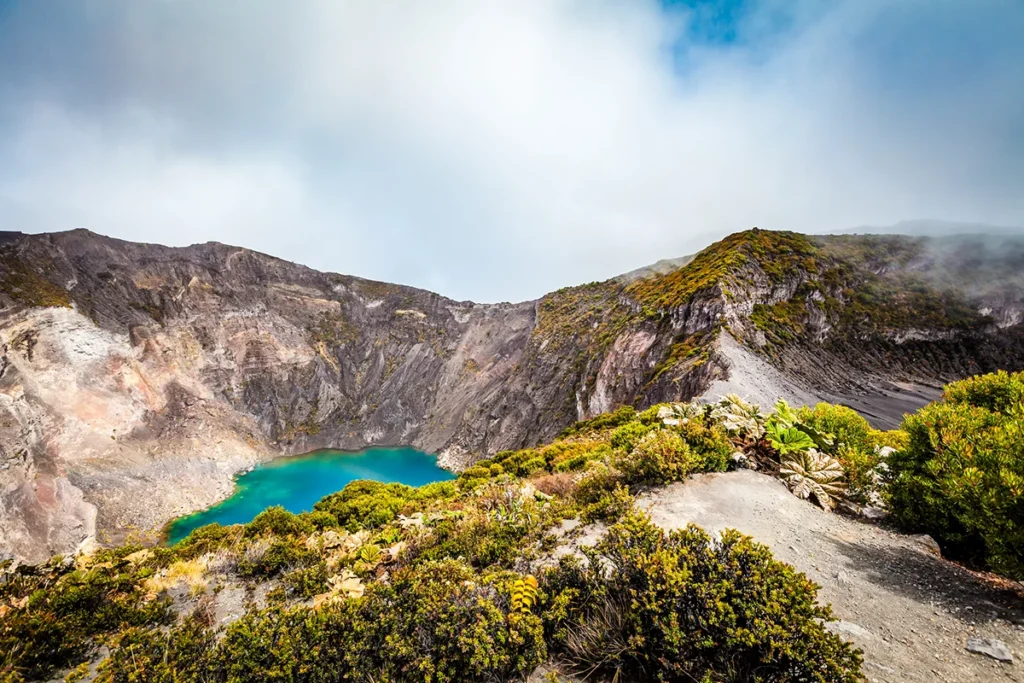 Der türkisfarbene Kratersee des Vulkans Irazú in Costa Rica liegt inmitten einer kargen Vulkanlandschaft. Die steilen Kraterwände umgeben das leuchtend blaue Wasser unter einem weiten Himmel.