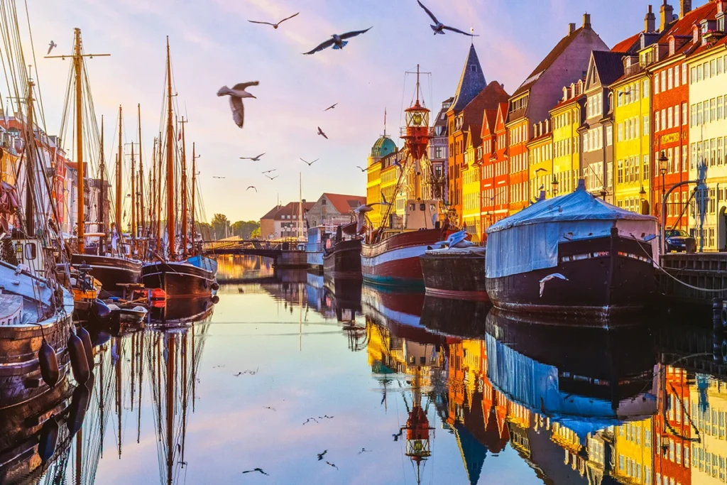 Der Hafen Nyhavn in Kopenhagen liegt in warmem Abendlicht der Golden Hour. Bunte Häuser spiegeln sich im Wasser, Boote liegen am Kai und Möwen fliegen über dem Hafen.