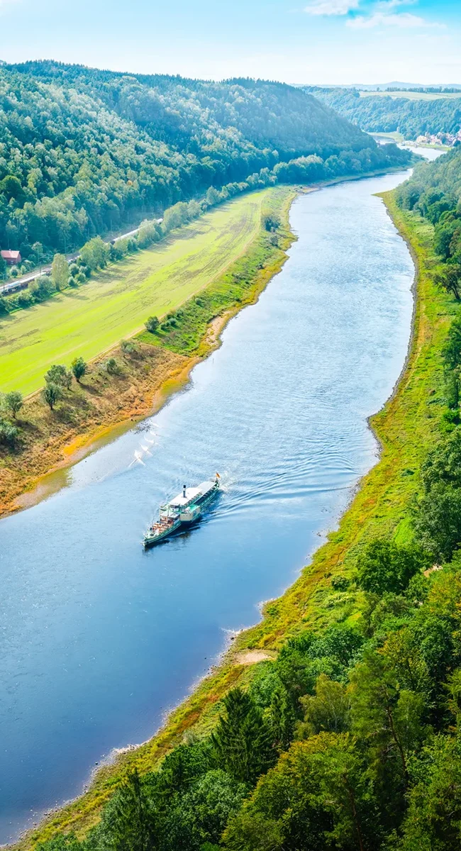 Ein Ausflugsschiff fährt an einem sonnigen Tag entlang der Elbe und vorbei an grünen Ufern.