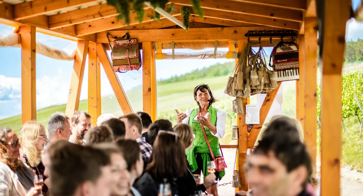 In einem gemütlichen, hölzernen Wagen moderiert eine Frau in grüner Tracht eine weinselige Gesellschaftsfahrt durch eine sonnige Weinberglandschaft in Hohenlohe.