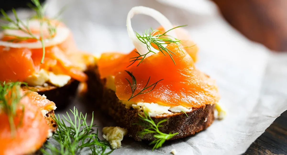 Ein kleines Fisch-Canapé mit Lachs, Dill und Zwiebeln liegt auf einem Stück Brot. Die Zutaten sind dekorativ auf dem Häppchen angerichtet.