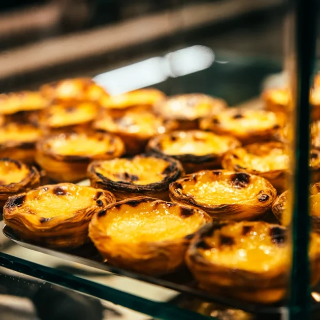 Mehrere Pastéis de Nata liegen in einer Auslage einer Bäckerei. Die kleinen portugiesischen Blätterteigtörtchen sind mit goldbrauner Vanillecreme gefüllt.