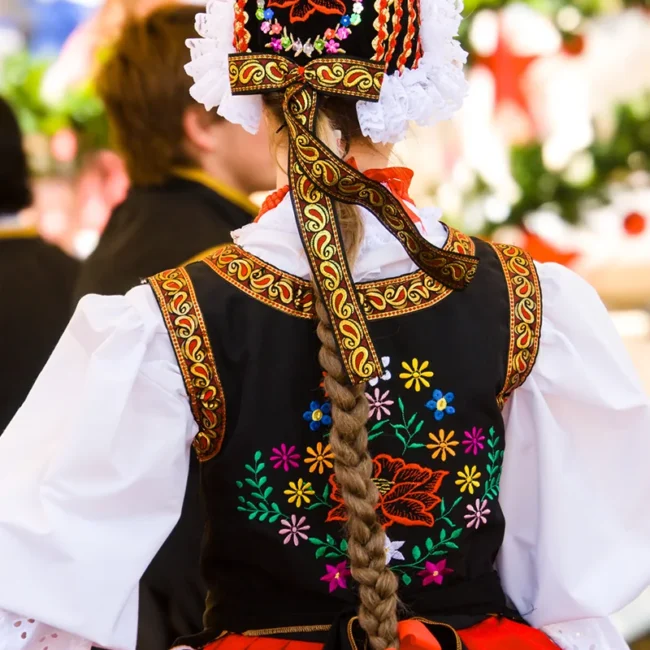 Eine Frau in traditioneller Folklorekleidung ist von hinten zu sehen. Sie trägt ein buntes Kleid mit bestickten Mustern und Bändern.