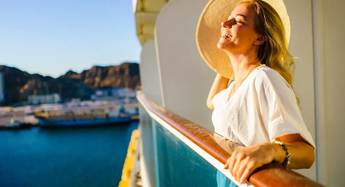 Eine Frau steht auf dem Balkon eines Kreuzfahrtschiffs und genießt die Sonne im Gesicht mit Blick auf das Meer.