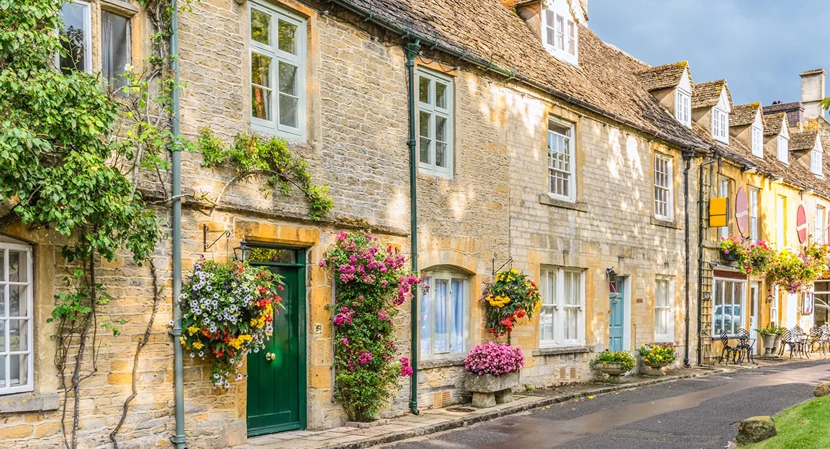 Eine charmante Häuserreihe im englischen Stow-on-the-Wold aus honigfarbenem Cotswold-Stein, geschmückt mit bunten Blumenampeln und üppig rankenden Kletterpflanzen.
