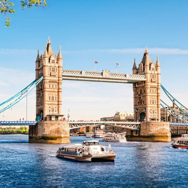 Die berühmte Londoner Tower Bridge in England bei strahlendem Sonnenschein über der Themse, während mehrere Ausflugsschiffe auf dem Wasser unterwegs sind.