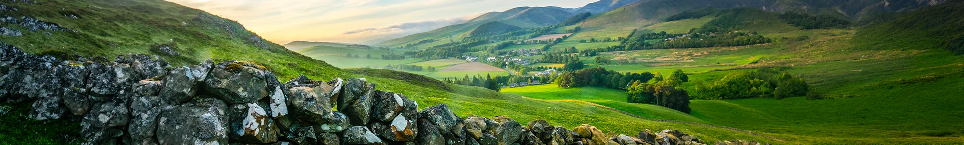 Eine uralte Steinmauer zieht sich im Vordergrund durch das saftig grüne Weideland in England, während im Hintergrund ein kleines Dorf friedlich im sonnendurchfluteten Tal liegt.