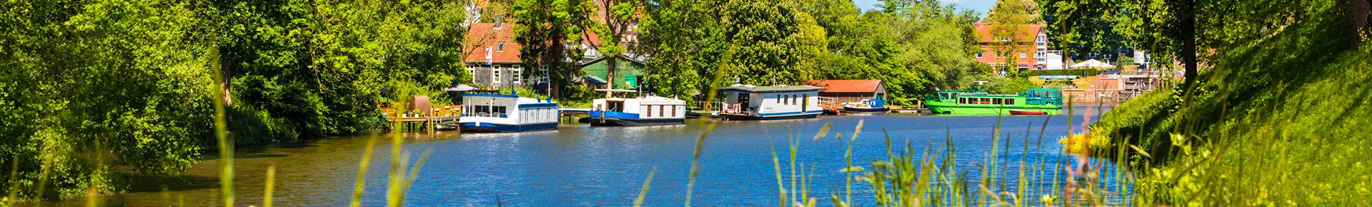 Eine idyllische Wasserlandschaft am Stader Burggraben mit mehreren modernen Hausbooten, die von dichtem Grün und historischen Gebäuden gesäumt werden.