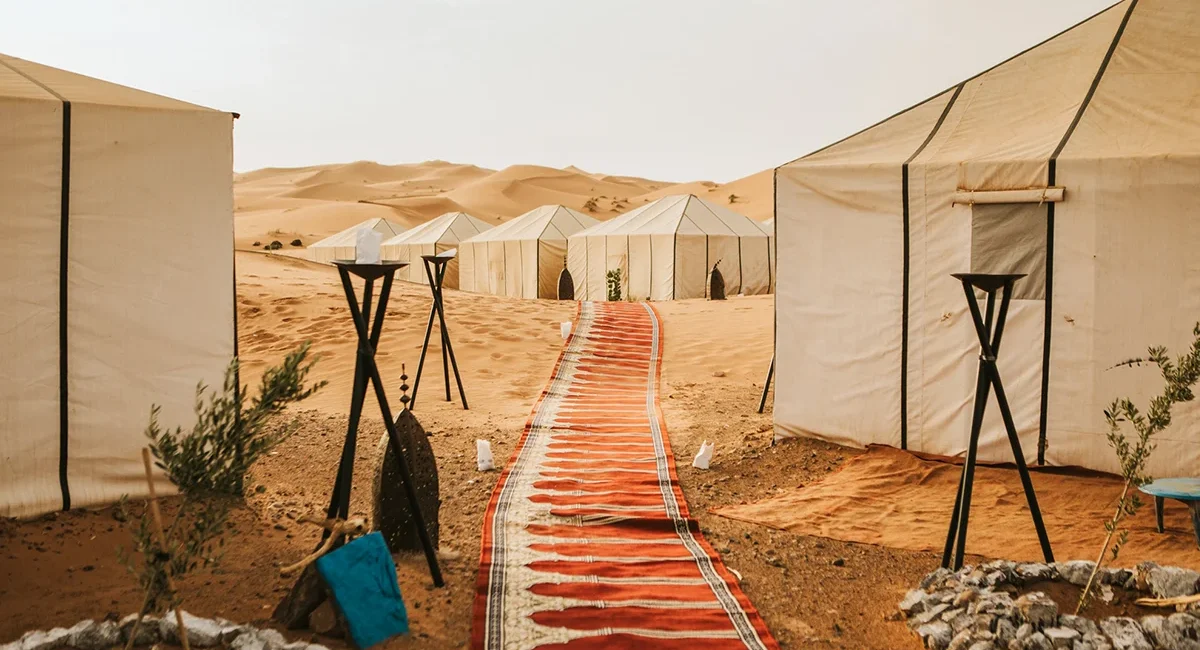 Ein luxuriöses Wüstencamp mit sandfarbenen Zelten und einem langen, roten Teppichläufer, das inmitten der sanften Dünen in Marokko zur Entspannung einlädt.
