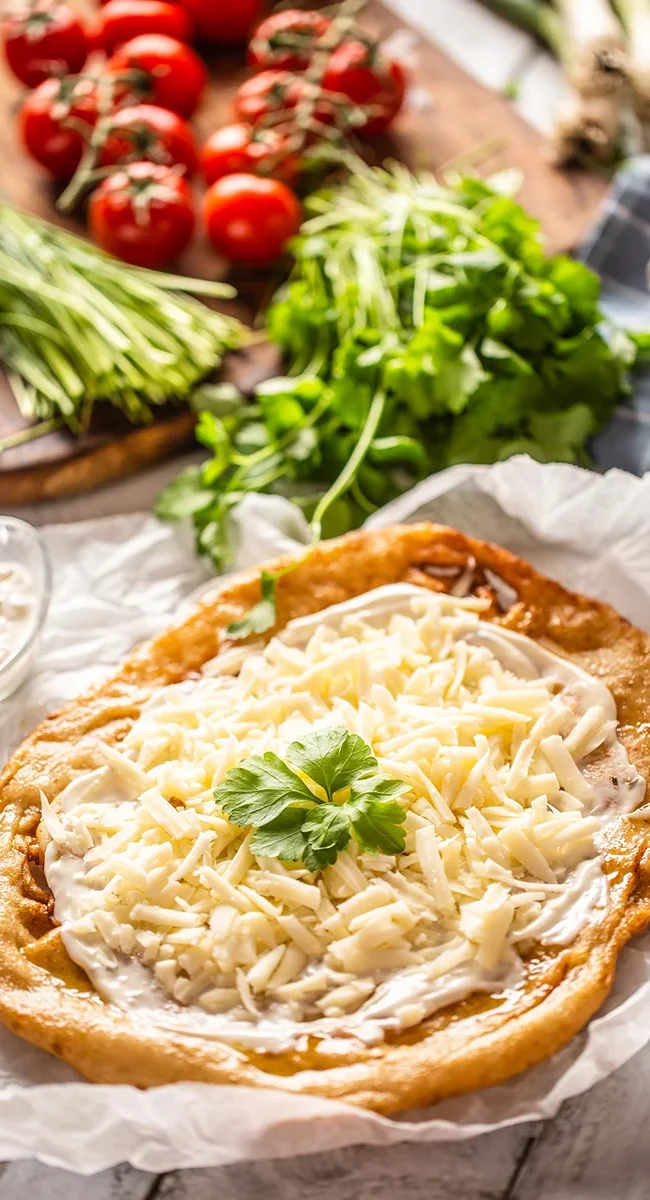Ein ungarisches Lángos, das auf Backpapier liegt und großzügig mit weißem Sauerrahm sowie einer dicken Schicht geriebenem Käse belegt ist.
