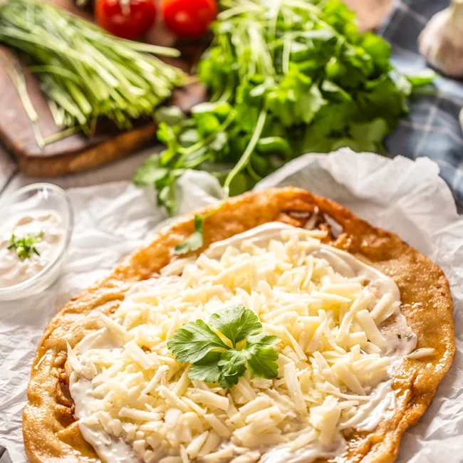 Ein ungarisches Lángos, das auf Backpapier liegt und großzügig mit weißem Sauerrahm sowie einer dicken Schicht geriebenem Käse belegt ist.