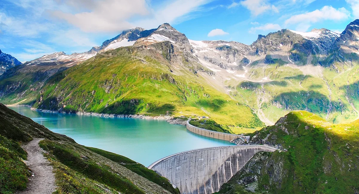 Eine Gebirgslandschaft mit zwei großen Staumauern und dem Wasserkraftwerk der Illwerke in Vorarlberg, die einen türkisblauen See inmitten grüner Hänge und schneebedeckter Gipfel einrahmen.